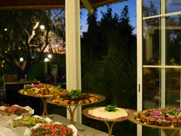 Catering buffet with salads and appetizers arranged in bowls and gold trays beside large glass windows at an evening event.