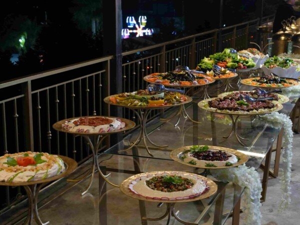 Outdoor evening catering buffet with assorted salads and appetizers displayed on gold trays along a terrace table.
