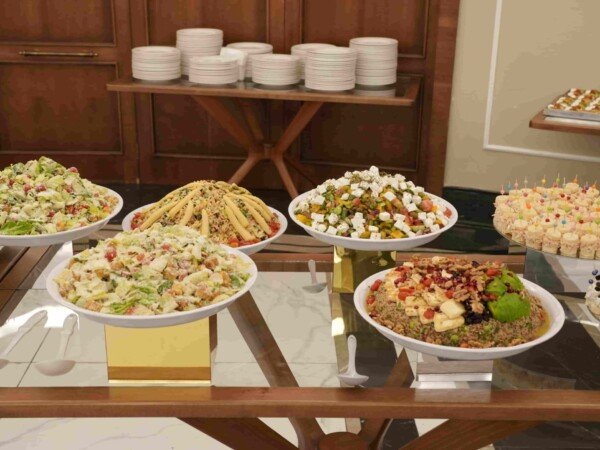 Indoor catering buffet with assorted salads, appetizers, and bite-sized sandwiches arranged on a glass table for an event.