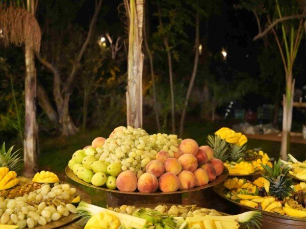 Outdoor fruit buffet with grapes, peaches, bananas, pineapple, and sliced fruit arranged on gold trays in a garden setting at night.