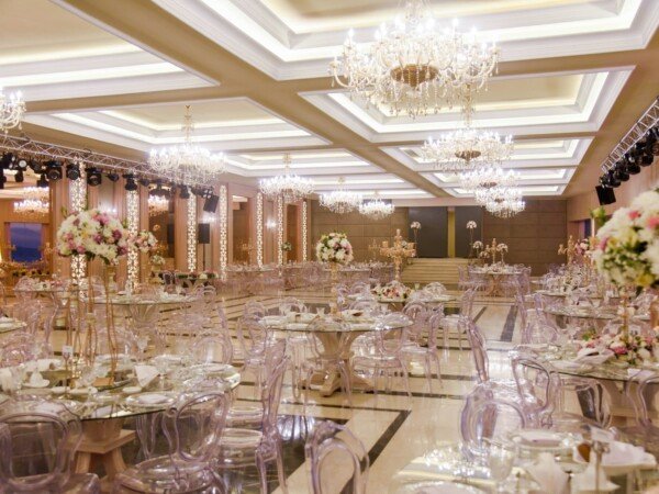Bright wedding ballroom with crystal chandeliers, clear chairs, glass tables, floral centerpieces, and warm elegant lighting.