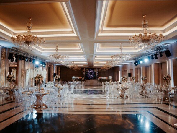 Spacious wedding ballroom with crystal chandeliers, clear chairs, round tables, floral centerpieces, and polished flooring.