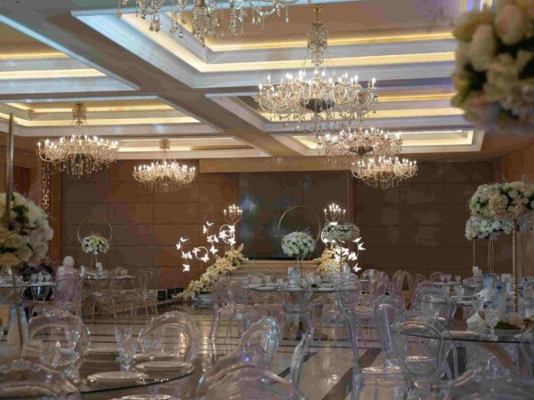 Elegant indoor wedding hall with crystal chandeliers, clear chairs, glass tables, and white floral centerpieces.