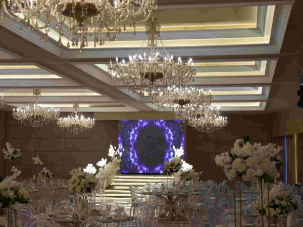 Indoor wedding reception hall with chandeliers, transparent chairs, floral centerpieces, and an illuminated blue stage backdrop.