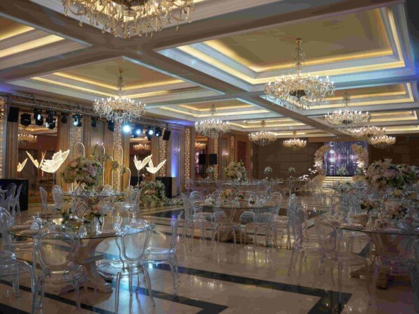 Elegant indoor wedding hall with crystal chandeliers, clear chairs, glass tables, floral centerpieces, and stage lighting