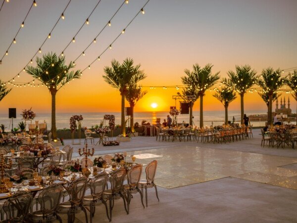 Seaside wedding reception at sunset with string lights, palm trees, clear chairs, decorated tables, and a glowing dance floor overlooking the ocean.