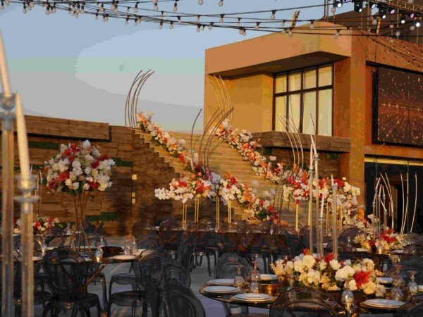 Outdoor wedding reception with black chairs, glass tables, floral arrangements, and string lights above the dining area at dusk.