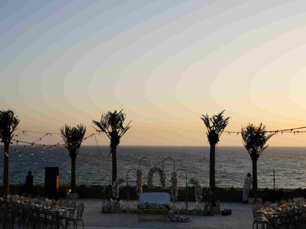 Seaside wedding venue at sunset with a glass dance floor, palm trees, string lights, and a floral ceremony setup overlooking the ocean.