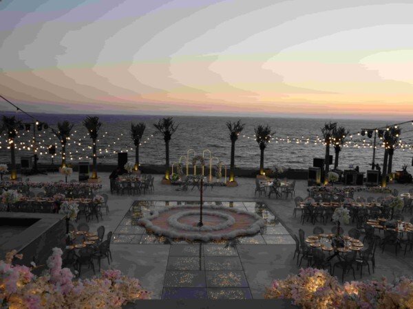 Seaside wedding reception at dusk with a glowing glass dance floor, round dining tables, palm trees, string lights, floral décor, and an ocean view.