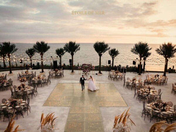 Seaside wedding reception at sunset with a bride and groom on a glowing dance floor, surrounded by round tables, palm trees, string lights, and ocean views.