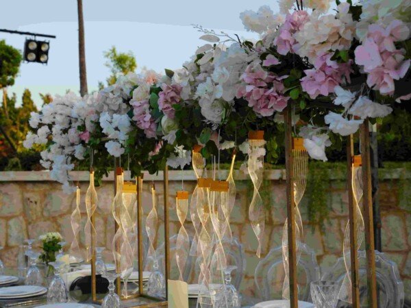 Outdoor wedding table setup with glass tables, clear chairs, floral overhead centerpieces, and elegant place settings in a garden venue.