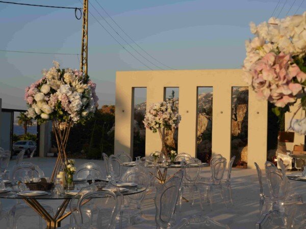 Outdoor wedding reception setup with clear chairs, glass tables, and tall floral centerpieces at sunset.