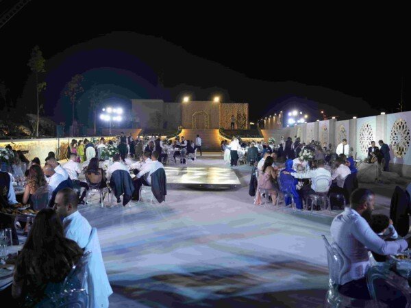 Outdoor nighttime event reception with guests seated at round tables around an illuminated dance floor, decorative wall panels, and bright stage lighting.