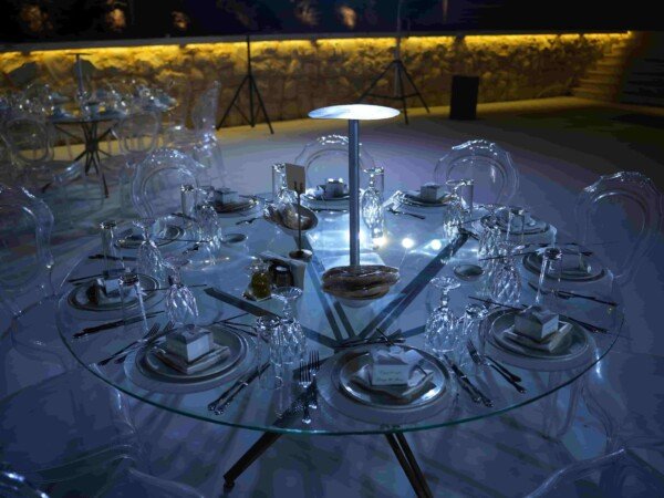 Elegant outdoor event table setup with glass dining table, clear chairs, place settings, glassware, and soft blue evening lighting.