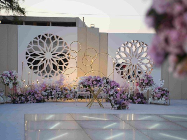 Elegant outdoor wedding stage with floral arrangements, decorative white backdrop panels, gold circle accents, candles, and glass chairs at dusk.