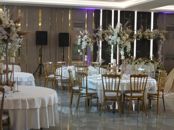 Indoor wedding reception hall with round tables, gold chairs, white floral centerpieces, and an elegant sweetheart table with a decorative harp.