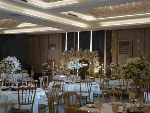 Elegant indoor wedding reception hall with round tables, gold chairs, floral centerpieces, and soft chandelier lighting.