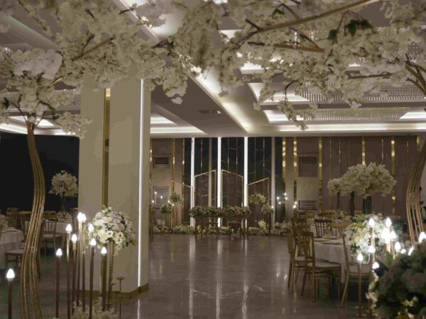 Indoor wedding hall with white floral aisle decorations, hanging ceiling flowers, soft lighting, and elegant reception tables.