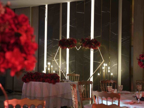 Indoor event reception with round dining tables, red floral arrangements, candlelit décor, and an elegant sweetheart table backdrop.